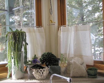 May include: A window with white curtains and a variety of potted plants, including a succulent, a violet, and a fern. There is a small bird figurine hanging from the ceiling.