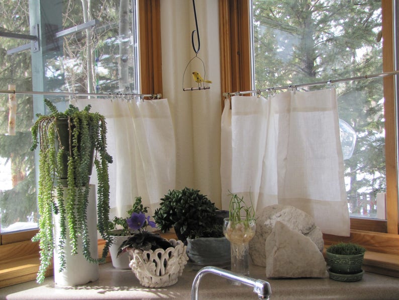 custom classic linen curtains for sale. shown in photo is A window with white curtains and a variety of potted plants, including a succulent, a violet, and a small tree. There is a bird on a swing hanging from the ceiling.