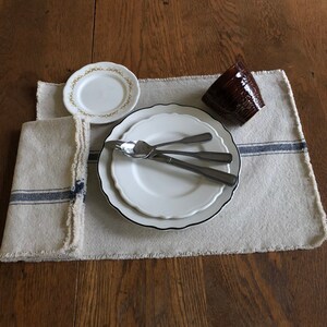 May include: A white dinner plate with a black rim and a smaller white plate with a gold rim on top. Two silver forks and a silver spoon are on the plates. A brown ceramic mug is on a white linen placemat with blue stripes.