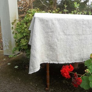 May include: A white linen tablecloth with a frayed edge, draped over a wooden table. The tablecloth is in a natural setting with green foliage and red flowers in the background.