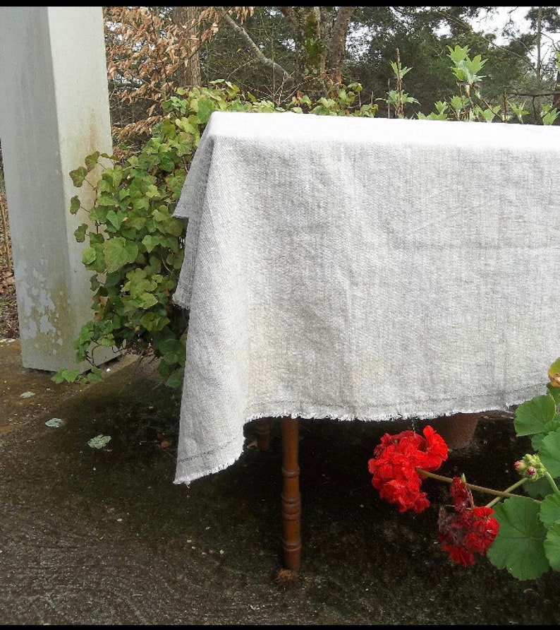 May include: A white linen tablecloth with a frayed edge, draped over a wooden table. The tablecloth is in a natural setting with green foliage and red flowers in the background.