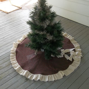May include: A red and green plaid Christmas tree skirt with a white ruffled edge. The skirt is made of burlap and has a bow on the side.