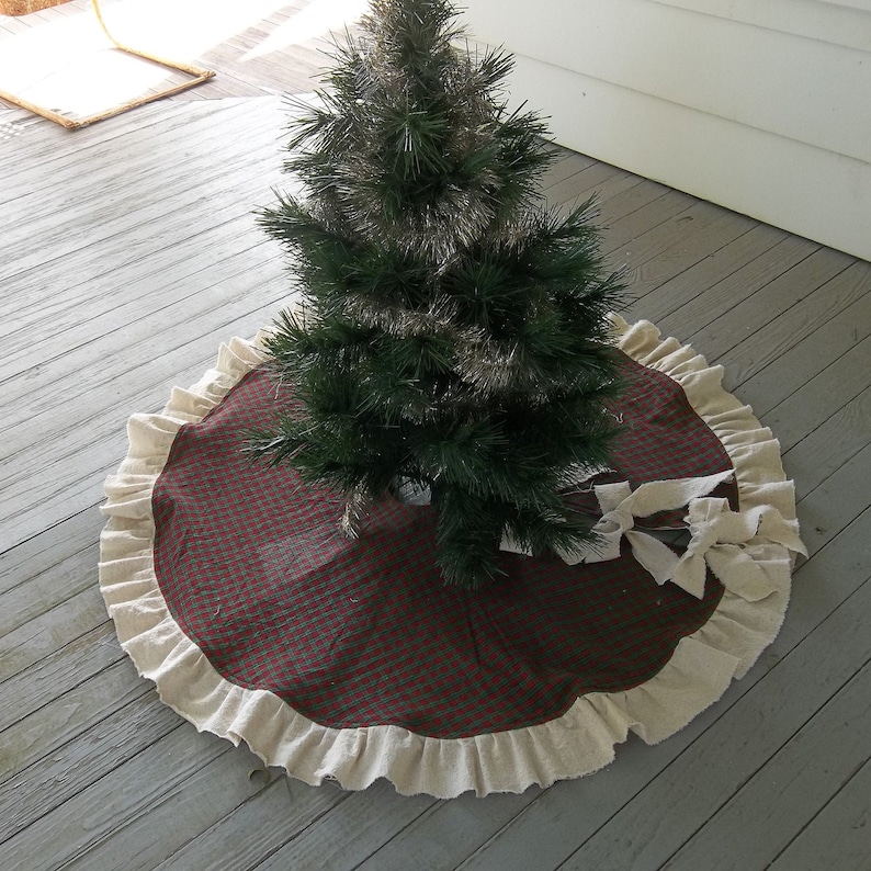 May include: A red and green plaid Christmas tree skirt with a white ruffled edge. The skirt is made of burlap and has a bow on the side.