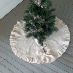 May include: A white, ruffled Christmas tree skirt with a large bow in the center. The skirt is made of a soft, natural fabric and has a simple, elegant design.