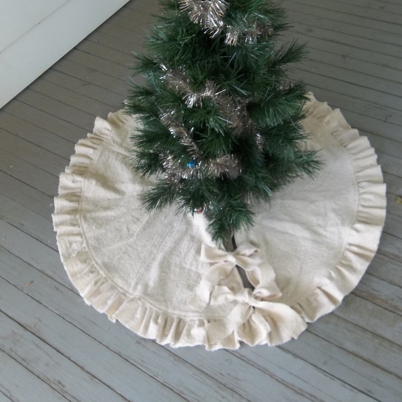 May include: A white, ruffled Christmas tree skirt with a large bow in the center. The skirt is made of a soft, natural fabric and has a simple, elegant design.