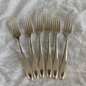 May include: Six silver forks with ornate handles. The forks are arranged in a row on a white fabric background.