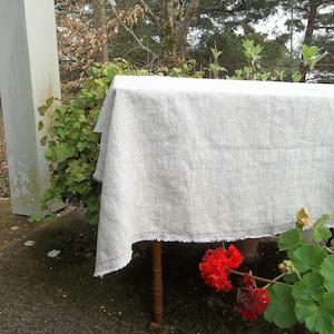 May include: A white linen tablecloth with a frayed edge, draped over a wooden table outdoors. The tablecloth is in a natural setting with green foliage and red flowers in the foreground.