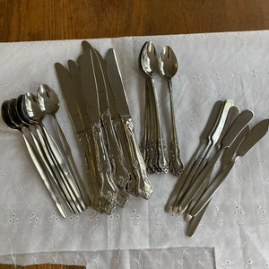 May include: A set of silverware, including knives, forks, and spoons, with ornate handles. The silverware is arranged on a white tablecloth with a delicate floral pattern.