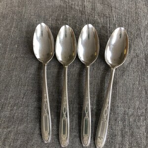 May include: Four silver spoons with ornate designs on the handles. The spoons are laid out in a row on a gray fabric surface.