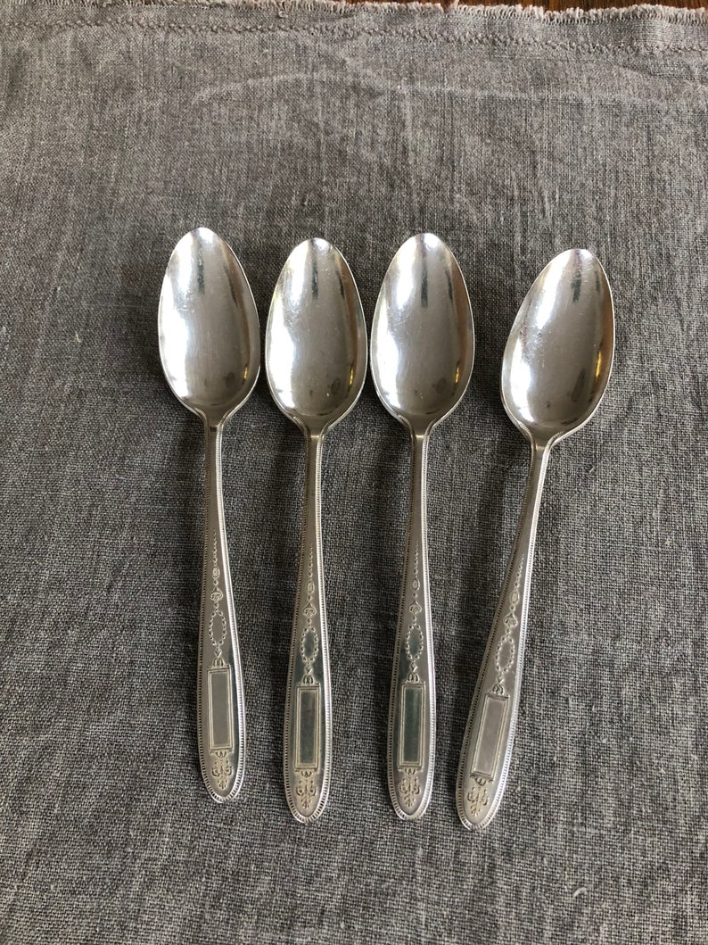 May include: Four silver spoons with ornate designs on the handles. The spoons are laid out in a row on a gray fabric surface.