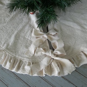 May include: A white cotton Christmas tree skirt with a ruffled edge and two large fabric bows tied in the center.