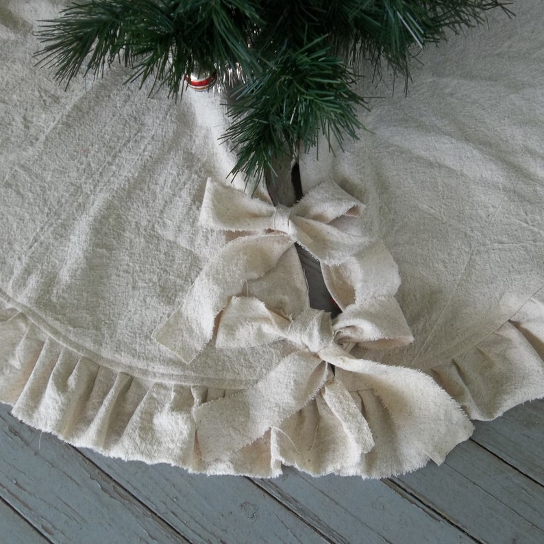 May include: A white cotton Christmas tree skirt with a ruffled edge and two large fabric bows tied in the center.