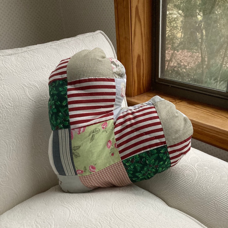 May include: A heart-shaped patchwork pillow with red, green, and white fabric. The pillow is made of different patterns and textures, including stripes, floral prints, and plaid.