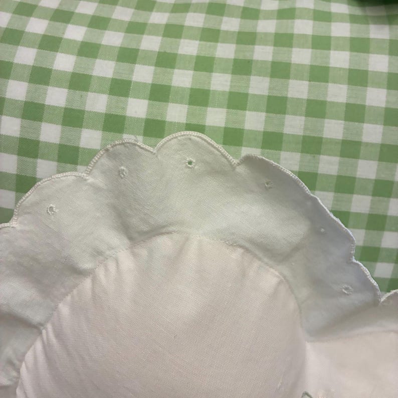 May include: A white fabric pillow featuring a scalloped edge with small embroidered eyelet details. The pillow is positioned against a background of light green and white gingham fabric, creating a contrast between the solid white and pattern.