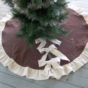 May include: A red and green plaid Christmas tree skirt with a white ruffled edge and three white fabric bows.