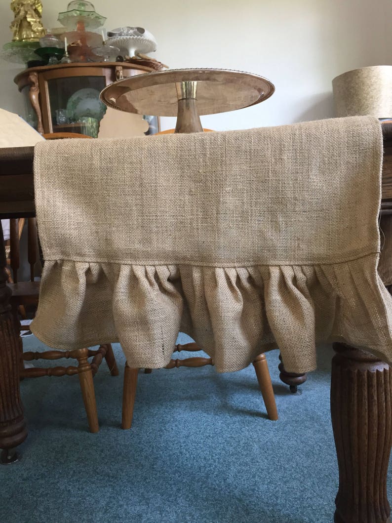 May include: A brown burlap table runner with a ruffled edge. The runner is draped over a wooden table and chairs.