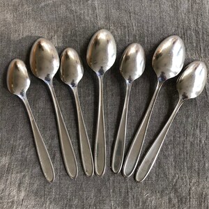 May include: A set of eight silver spoons arranged in a semi-circle on a gray fabric surface.