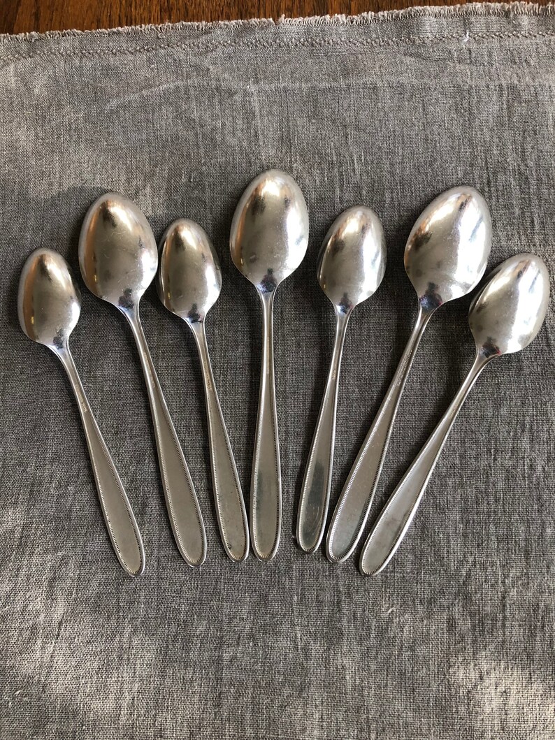 May include: A set of eight silver spoons arranged in a semi-circle on a gray fabric surface.
