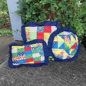 May include: Three handmade patchwork pillows in various shapes. The pillows feature a mix of colorful fabric squares and floral patterns, framed by a dark blue ruffled border. The pillows are arranged on a concrete surface with greenery in the background.