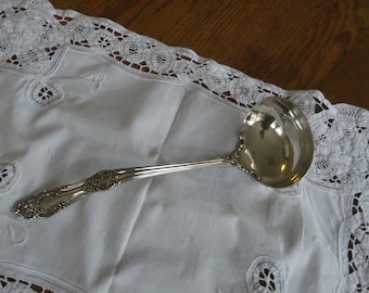 May include: A silver ladle with an ornate handle, resting on a white lace tablecloth.