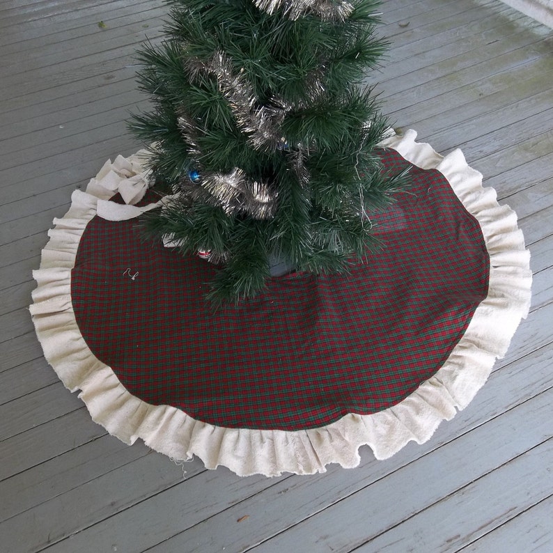 May include: A red and green plaid Christmas tree skirt with a white ruffled edge. The skirt is made of fabric and has a simple design.