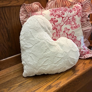May include: A white heart-shaped pillow with a textured surface, resting on a wooden bench. Behind it, a decorative pillow with a red and white toile pattern and a ruffled edge.