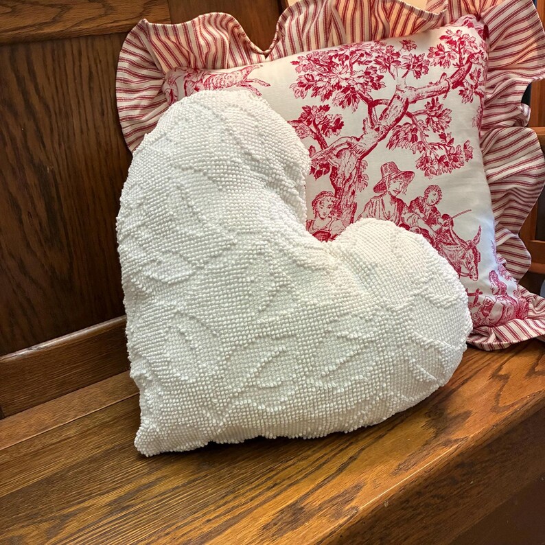 May include: A white heart-shaped pillow with a textured surface, resting on a wooden bench. Behind it, a decorative pillow with a red and white toile pattern and a ruffled edge.