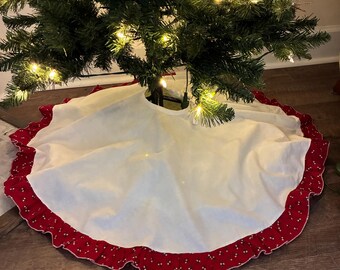 May include: A white Christmas tree skirt with a red patterned border. The skirt is circular and sits beneath a decorated Christmas tree with lights. The border has a repeating pattern of small white dots on a red background.