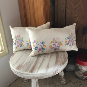 May include: Two embroidered throw pillows with floral designs in shades of pink, blue, yellow, and green on a white background. The pillows are sitting on a white painted wooden stool.