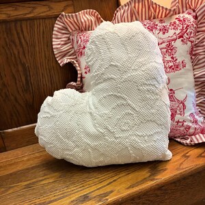 May include: A large, textured white heart-shaped pillow with a raised, dotted pattern. The pillow is resting on a wooden surface, with two decorative pillows in the background. One pillow has a red and white striped pattern, and the other has a red toile design.