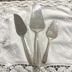 Może przedstawiać: Zestaw trzech srebrzystych przyborów do serwowania, w tym łopatka do ciasta z gładką krawędzią, duża płaska łopatka z ząbkowaną krawędzią oraz mniejsza ozdobna łopatka z wytłoczonym wzorem kwiatowym i kratką.