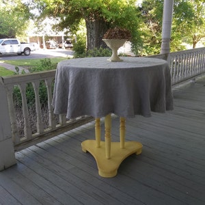 May include: A gray linen tablecloth draped over a yellow wooden table with a white pedestal bowl on top. The table is on a wooden porch with white railings.