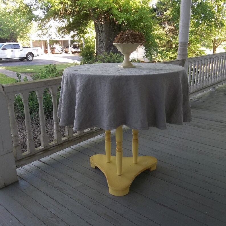May include: A gray linen tablecloth draped over a yellow wooden table with a white pedestal bowl on top. The table is on a wooden porch with white railings.
