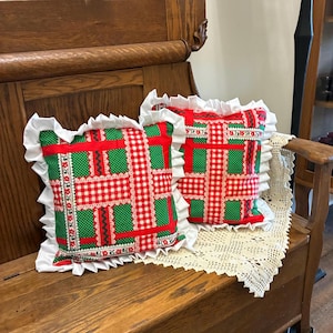 May include: Two square throw pillows featuring a patchwork design of red and white gingham, green polka dot fabric, and floral trim accents. Each pillow is finished with a white ruffled fabric border along all four edges of the square shape.