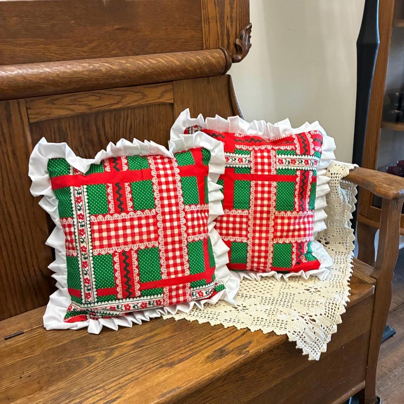 May include: Two square throw pillows featuring a patchwork design of red and white gingham, green polka dot fabric, and floral trim accents. Each pillow is finished with a white ruffled fabric border along all four edges of the square shape.
