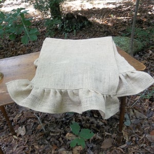 May include: A natural burlap table runner with a ruffled edge. The runner is laying on a wooden table in a forest setting.