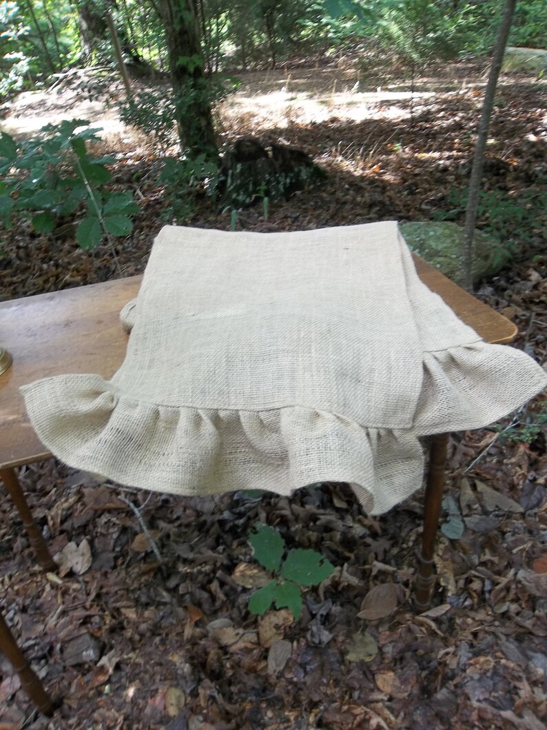 May include: A natural burlap table runner with a ruffled edge. The runner is laying on a wooden table in a forest setting.