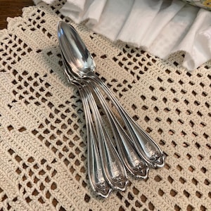May include: Set of five silver spoons with ornate handles. The spoons are arranged in a fan shape on a crocheted doily.