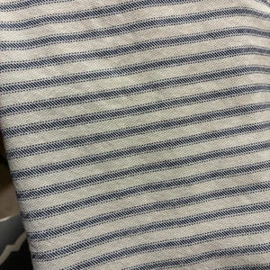 Op de afbeelding: Close-up van een witte stof met gelijkmatig verdeelde, horizontale marineblauwe strepen. De stof heeft een getextureerd oppervlak, waarbij de strepen op de witte achtergrond lijken te zijn gestikt. Het ontwerp heeft een klassieke, nautische uitstraling.