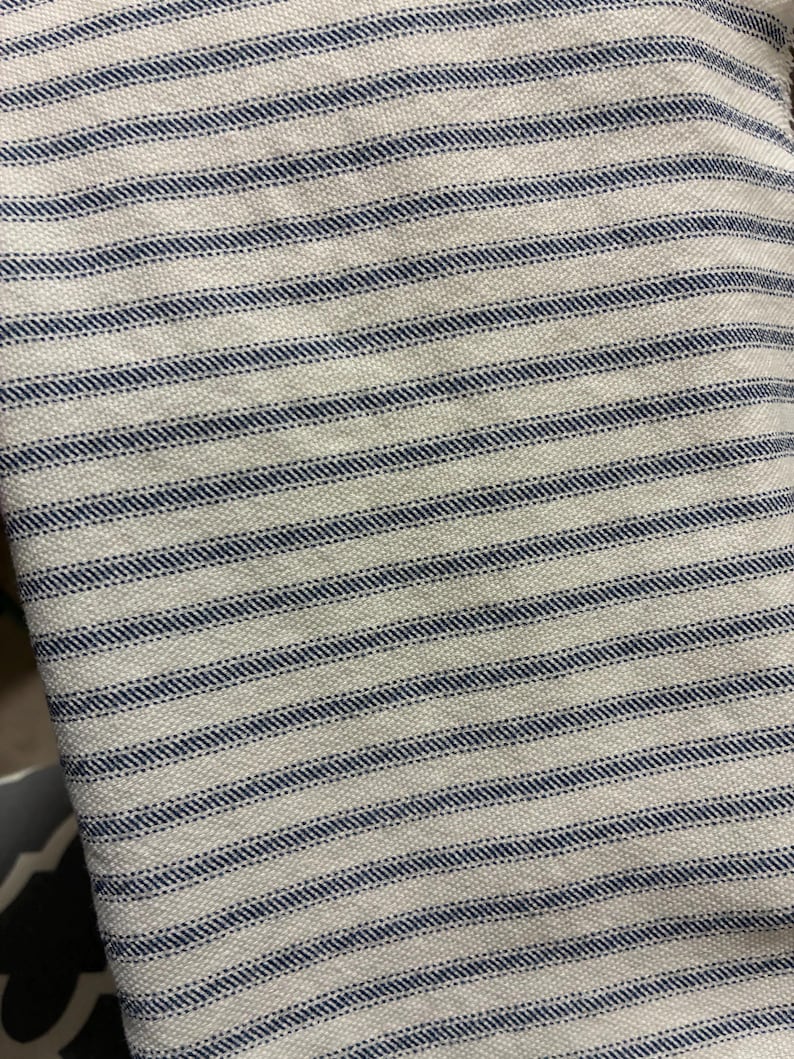 Op de afbeelding: Close-up van een witte stof met gelijkmatig verdeelde, horizontale marineblauwe strepen. De stof heeft een getextureerd oppervlak, waarbij de strepen op de witte achtergrond lijken te zijn gestikt. Het ontwerp heeft een klassieke, nautische uitstraling.