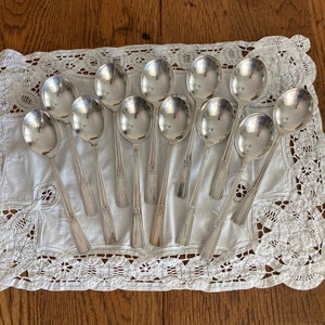 May include: A set of 12 silver spoons with ornate handles, arranged on a white lace tablecloth.