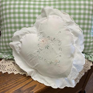 May include: A white heart-shaped pillow featuring a scalloped edge with eyelet detailing. The center of the pillow is decorated with delicate floral embroidery in soft shades of pink and green, showcasing intricate stitching and vine patterns.