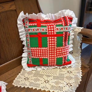 May include: A square throw pillow featuring a patchwork pattern of red and white gingham, green polka dots, and floral borders. The pillow is finished with a wide, white ruffled trim along all four edges, creating a decorative border.