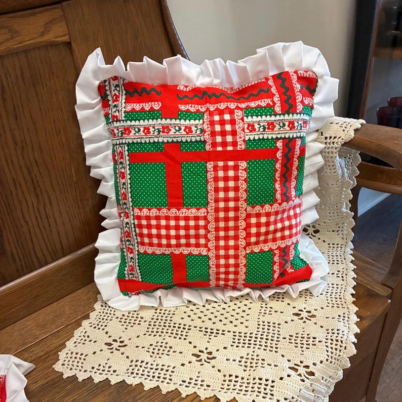 May include: A square throw pillow featuring a patchwork pattern of red and white gingham, green polka dots, and floral borders. The pillow is finished with a wide, white ruffled trim along all four edges, creating a decorative border.