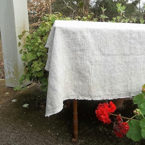 May include: A white linen tablecloth with a frayed edge, draped over a wooden table. The tablecloth is slightly wrinkled and has a subtle texture.