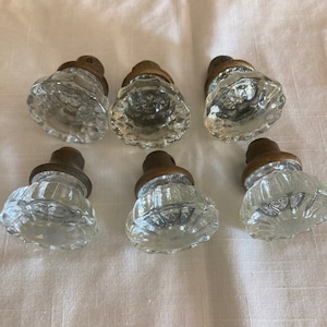 May include: A set of six clear glass door knobs with faceted surfaces and circular patterns. Each knob features a metallic bronze-toned base with a cylindrical neck, providing a contrast to the transparent, textured glass heads.