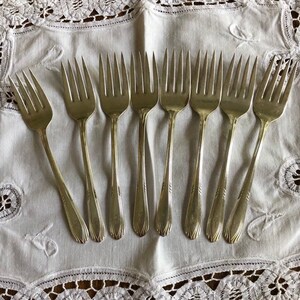 May include: Ten silver forks laid out on a white lace tablecloth. The forks have a simple design with a rounded handle and a slightly curved tines.
