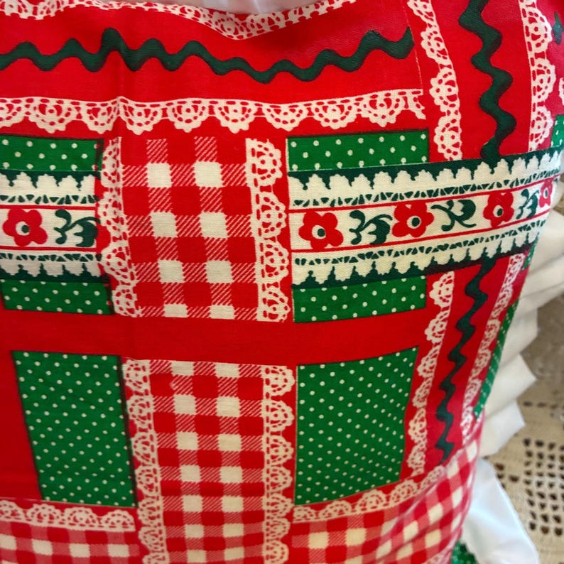 May include: A fabric pillow cover featuring a patchwork design with red and white gingham squares, solid red sections, and green panels with white polka dots. The pattern is accented by white lace-like trim and dark green wavy lines.