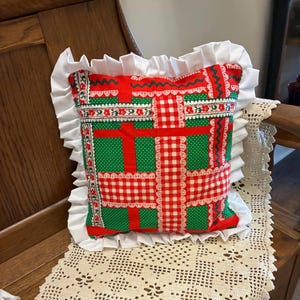 May include: A square throw pillow featuring a patchwork-style pattern of red and green fabric with white lace trim accents. The pillow is bordered by a wide, gathered white fabric ruffle along all four edges, creating a decorative frame.