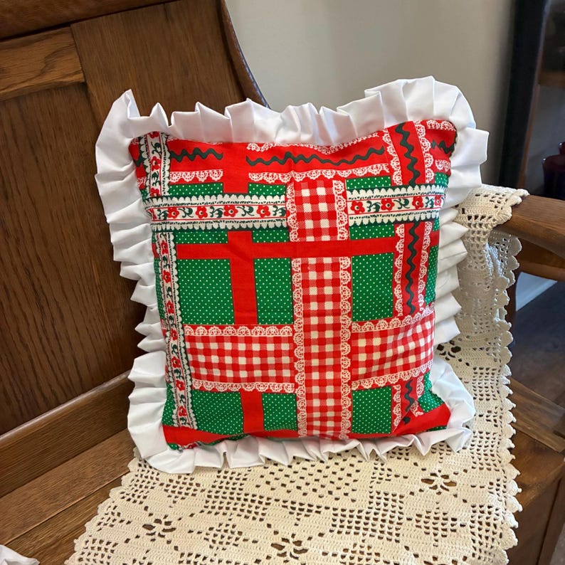 May include: A square throw pillow featuring a patchwork-style pattern of red and green fabric with white lace trim accents. The pillow is bordered by a wide, gathered white fabric ruffle along all four edges, creating a decorative frame.
